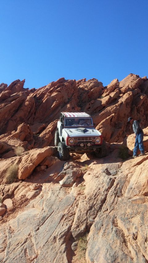 1979 Ford Bronco - A white and red 1979 Ford Bronco is parked on a rocky, off-road trail with a person standing nearby. The Bronco has large off-road tires an…