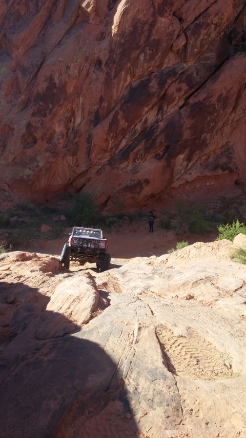 1979 Ford Bronco - A red and white 1979 Ford Bronco is navigating a rocky, off-road trail with a large red rock formation in the background. A person stands i…