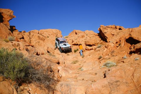 1979 Ford Bronco - A white Ford Bronco with an American flag decal on the roof navigates a steep, rocky, off-road trail under a clear blue sky. A person in an…