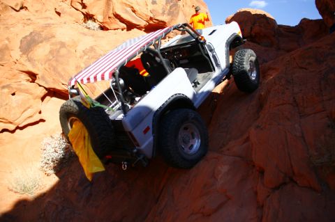 1979 Ford Bronco - A white Ford Bronco is navigating a steep, rocky incline in a desert landscape. The vehicle has a striped canopy over the open-top cabin an…