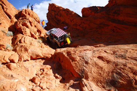 1979 Ford Bronco - A modified off-road vehicle with an American flag draped over its roof rack navigates a steep, rocky, orange desert terrain.