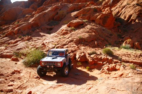 1979 Ford Bronco - A white and red classic Ford Bronco is shown driving off-road on a sandy trail surrounded by red rock formations. The vehicle is equipped w…