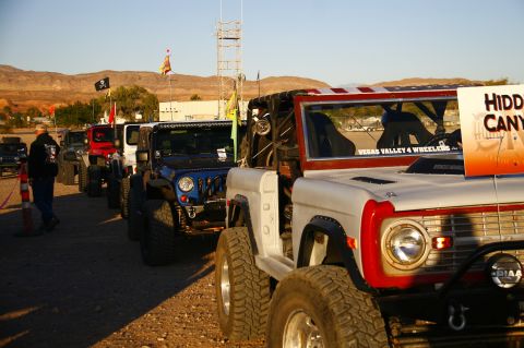 1979 Ford Bronco - A line of off-road vehicles, including a white and red Ford Bronco and a blue Jeep Wrangler, are parked on a dirt lot with mountains in the…