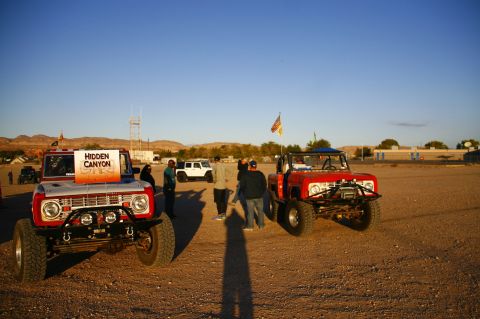 1979 Ford Bronco - Two modified Ford Broncos are parked on a dirt lot under a clear blue sky. A sign on the front Bronco reads "HIDDEN CANYON 750".