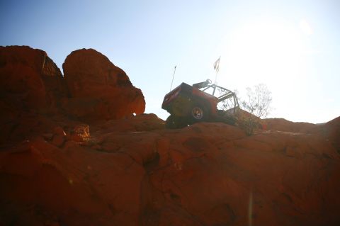 1979 Ford Bronco - An orange off-road vehicle with a roll cage is parked on a rocky, desert terrain under a bright, sunny sky. The vehicle has two flags attac…