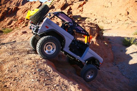 1979 Ford Bronco - A gray 1979 Ford Bronco is shown off-roading on a steep, rocky, desert terrain. The vehicle has a spare tire mounted on the rear and an Ame…