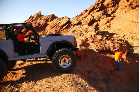 1979 Ford Bronco - A gray Ford Bronco is parked on a rocky, desert terrain with a driver in an orange shirt visible inside. Another person in an orange shirt…