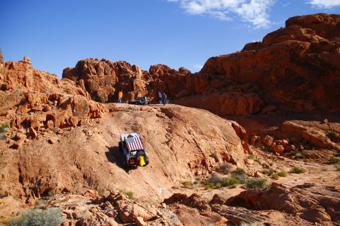 1979 Ford Bronco - A lifted, gray Ford Bronco with an American flag draped over its roof is parked on a rocky, desert hillside. Several people stand around ot…
