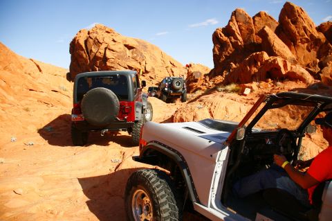 1979 Ford Bronco - A white open-top Ford Bronco is driving on a dusty orange trail in a desert landscape with red rock formations. Several other off-road vehi…