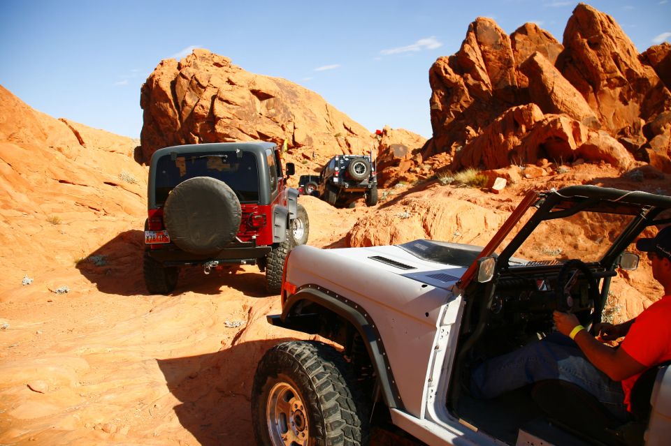 White open-top Ford Bronco driving on an off-road trail in a desert with red rocks.