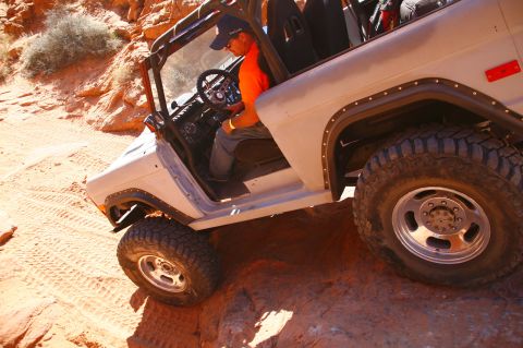 1979 Ford Bronco - A white Ford Bronco is navigating a rocky, sandy terrain with a driver at the wheel. The vehicle is angled downwards, with its large tires…