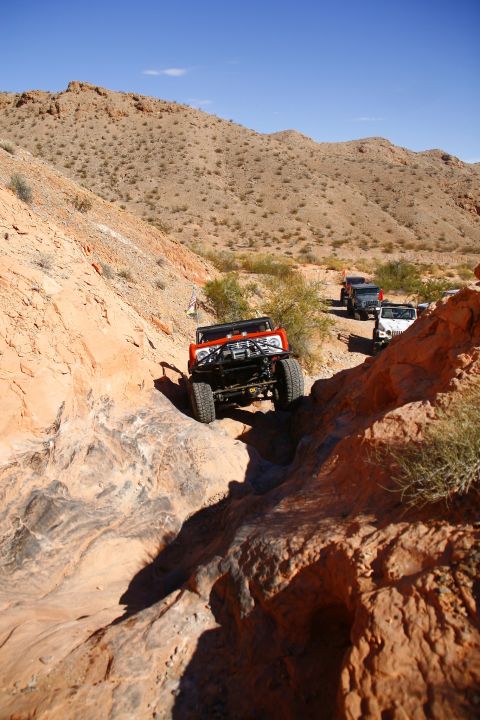 1979 Ford Bronco - An orange 1979 Ford Bronco is navigating a rocky off-road trail in a desert landscape. Several other Jeeps are visible further down the tra…