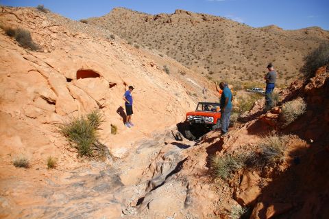 1979 Ford Bronco - An orange Ford Bronco is navigating a rocky, off-road trail with three people observing. The Bronco is positioned on a steep incline with r…