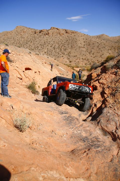 1979 Ford Bronco - An orange Ford Bronco is navigating a rocky, off-road trail in a desert landscape. Several people are standing around watching the vehicle.