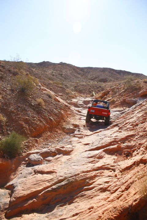1979 Ford Bronco - An orange Ford Bronco is driving up a rocky, off-road trail in a desert landscape under a bright, clear sky. The vehicle is viewed from the…