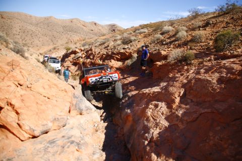 1979 Ford Bronco - An orange Ford Bronco is navigating a narrow, rocky canyon trail with a group of off-road vehicles behind it. A man stands on the side of t…