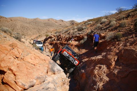 1979 Ford Bronco - An orange Jeep Wrangler is tilted precariously in a rocky canyon, with two people observing and another Jeep further back on the trail.