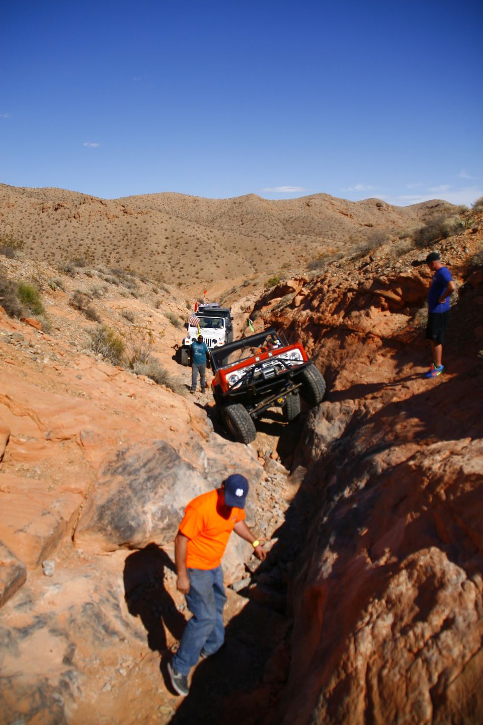 Orange 1979 Ford Bronco off-roading on a steep, rocky trail with other vehicles.