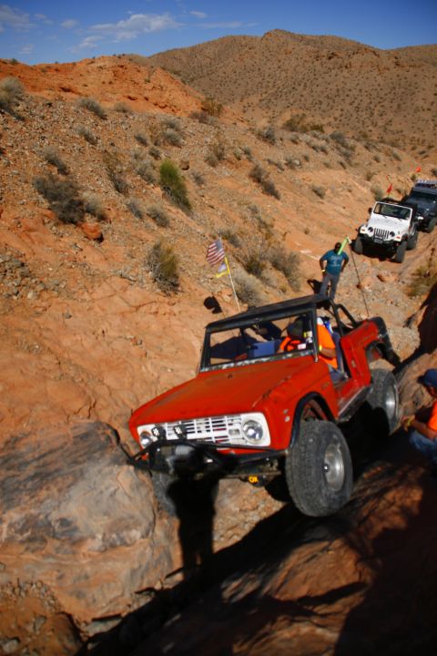 1979 Ford Bronco - An orange Ford Bronco is navigating a rocky, off-road trail with other Jeeps following behind. The Bronco is angled towards the right, with…
