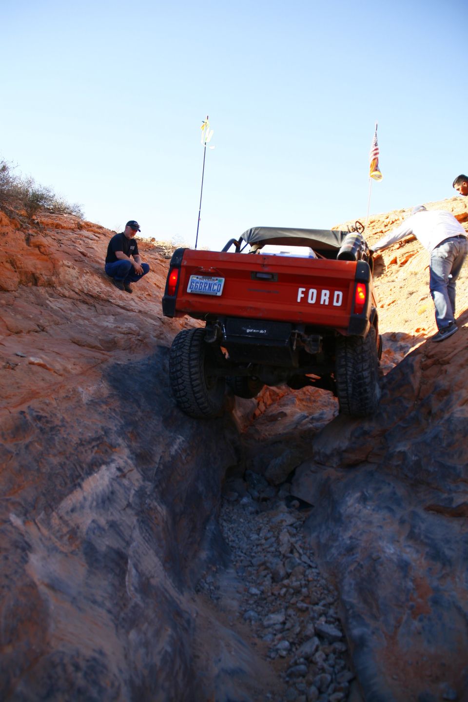Orange Ford Bronco driving down a rocky off-road trail with two people nearby.
