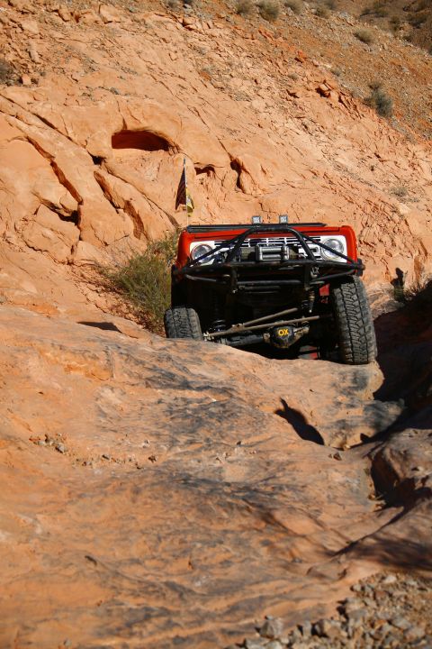 1979 Ford Bronco - A red 1979 Ford Bronco is navigating a rocky, off-road trail in a desert environment. The vehicle features a prominent aftermarket front bu…