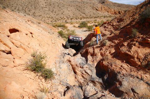 1979 Ford Bronco - A red 1979 Ford Bronco is navigating a steep, rocky canyon. A person in an orange shirt stands on the ridge above, watching the vehicle des…