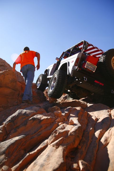 1979 Ford Bronco - A white off-road vehicle with an American flag-themed roll cage is climbing a rocky incline, with a person in an orange shirt walking behin…