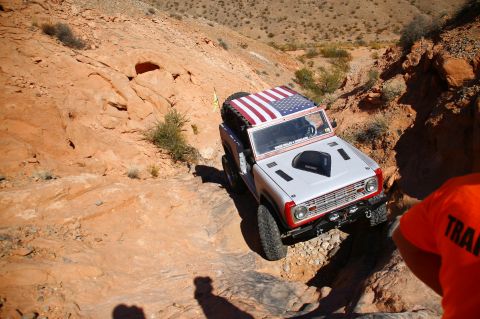 1979 Ford Bronco - A white Ford Bronco with an American flag decal on the roof is navigating a rocky, off-road trail in a desert environment. The vehicle is a…