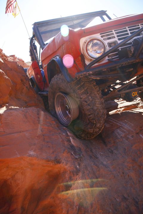 1979 Ford Bronco - A red Ford Bronco is navigating a rocky, off-road trail, with its front tire angled upwards on a large rock. The vehicle has a roll cage an…