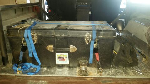 1979 Ford Bronco - A large black plastic trunk is secured with blue ratchet straps in the bed of a vehicle. A sticker on the trunk reads "DELUXE TRUNK".