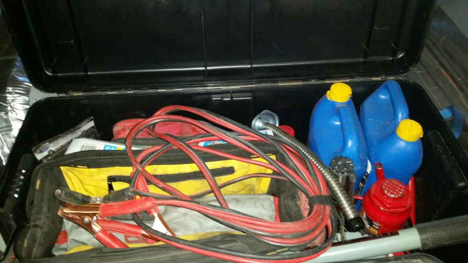 Open black storage box with jumper cables, fuel cans, and a jack.