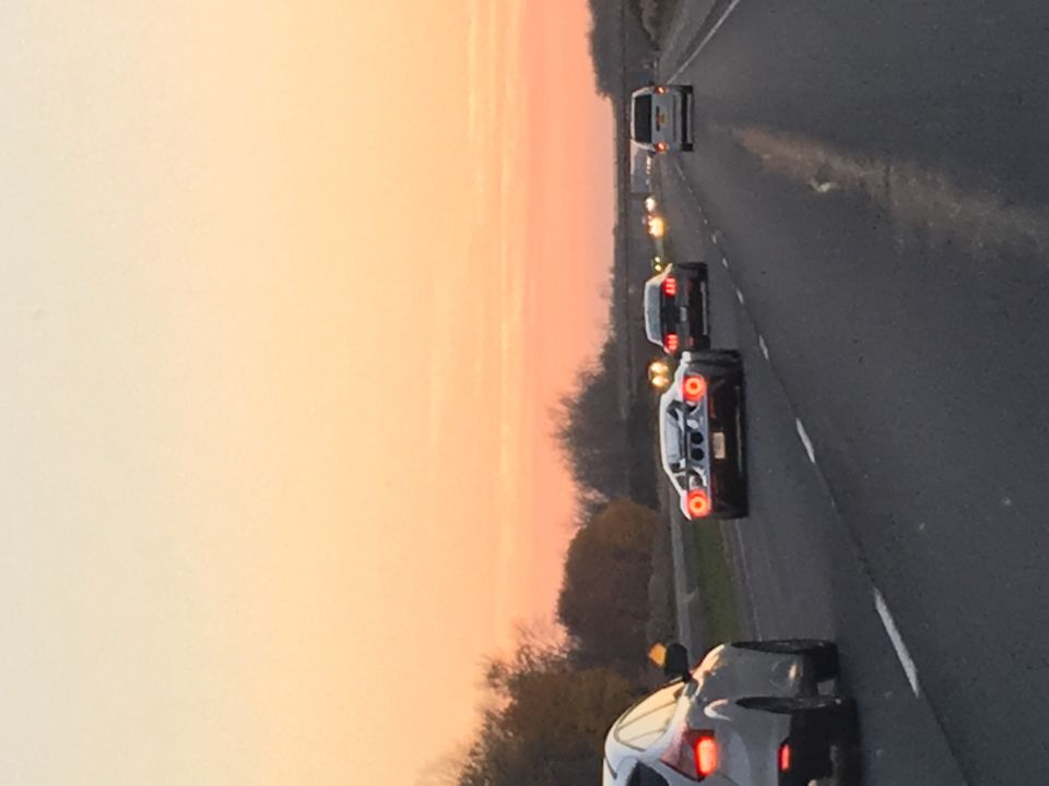 Line of traffic on a highway at sunset, dark SUV ahead with brake lights lit, orange sky.