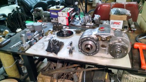 1985 Jeep CJ7 - A workbench is covered in automotive parts and tools, including a transfer case, air tools, gloves, and boxes of parts. A red car hood is v…