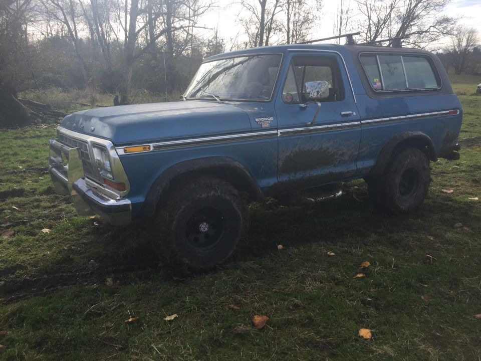 Blue Ford Bronco with chrome trim and roof rack, mud on body, parked on grassy field