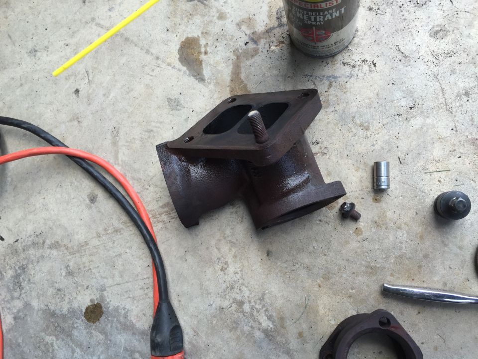 Rusty exhaust manifold part with bolts, wires, and a can of penetrant spray on a concrete surface