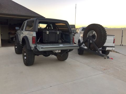 1988 Ford Bronco - A silver 1988 Ford Bronco is seen from the rear, with its tailgate down and a storage box inside, while towing a white pickup truck.