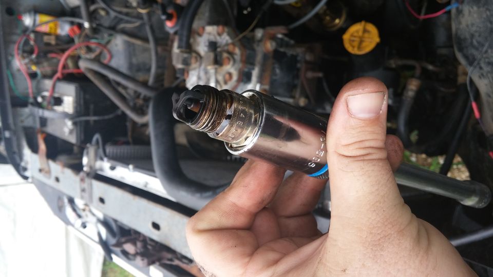 Hand holding a used spark plug with visible wear and carbon buildup. Engine components are blurred in the background