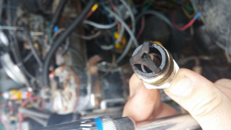 Hand holding a used spark plug with blackened electrodes, engine bay in background