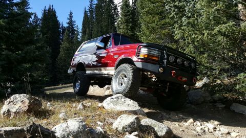 1996 Ford Bronco - A red 1996 Ford Bronco tackles a challenging off-road path through a wooded, rocky terrain.