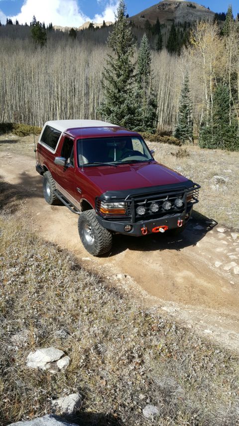 1996 Ford Bronco - A red 1996 Ford Bronco with a white top navigates a dirt road through a wooded mountain landscape.