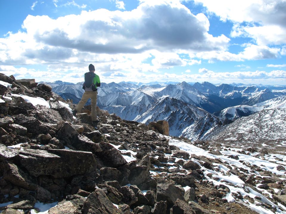 Hiker on rocky mountain peak overlooking snow-covered ranges under a cloudy sky