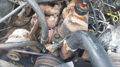 1984 Ford Bronco - Close-up view of a vehicle's engine bay, showing various hoses, wires, and rusty metal components. Text on a hose is visible.