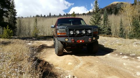 1996 Ford Bronco - A modified Ford Bronco is parked on a dirt road with a forest and mountain in the background.