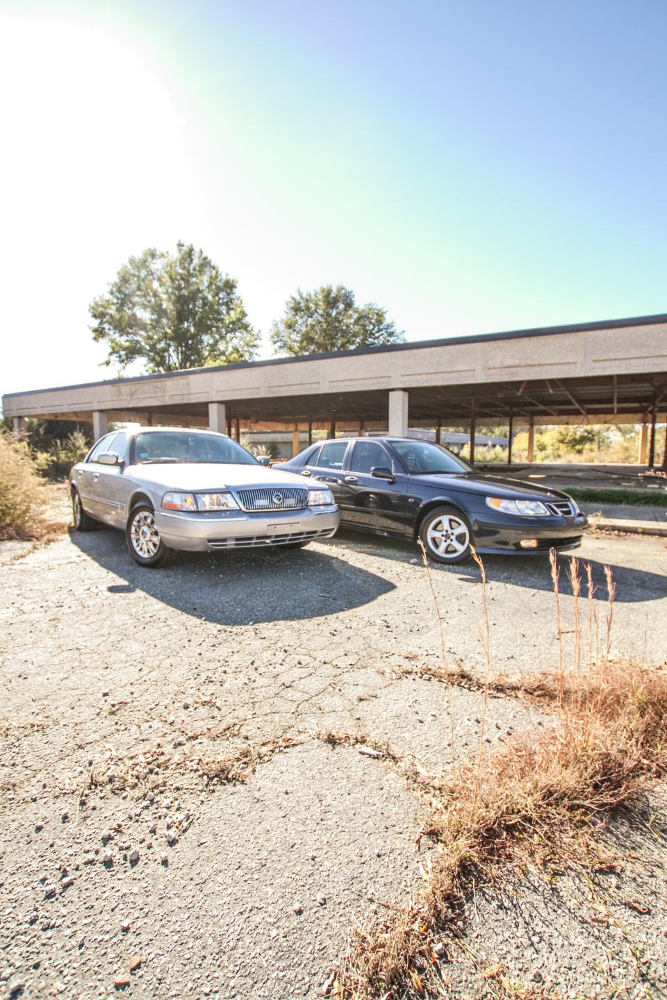 Silver Mercury Grand Marquis parked next to a dark blue Saab sedan in a weedy abandoned parking lot