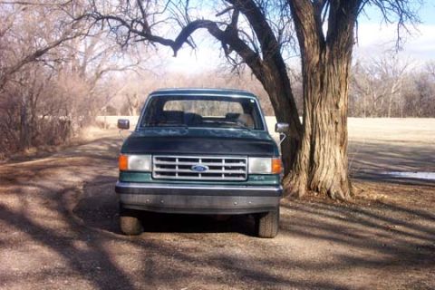1987 Ford Bronco - A dark green Ford F-Series truck is parked on a dirt road, with a large tree to its right.