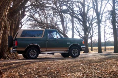 1987 Ford Bronco - A green Ford Bronco with a tan top is parked on a dirt road, surrounded by trees in the fall.