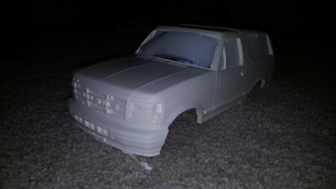 1996 Ford Bronco - A 3/4 front view of a white, unpainted Ford Bronco body shell with no wheels or tires is shown sitting on a gravel surface in a dark settin…