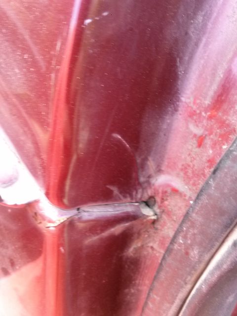 1996 Ford Bronco - A close-up view shows damage and rust on the red painted metal body of a vehicle, near a seam and a curved edge.