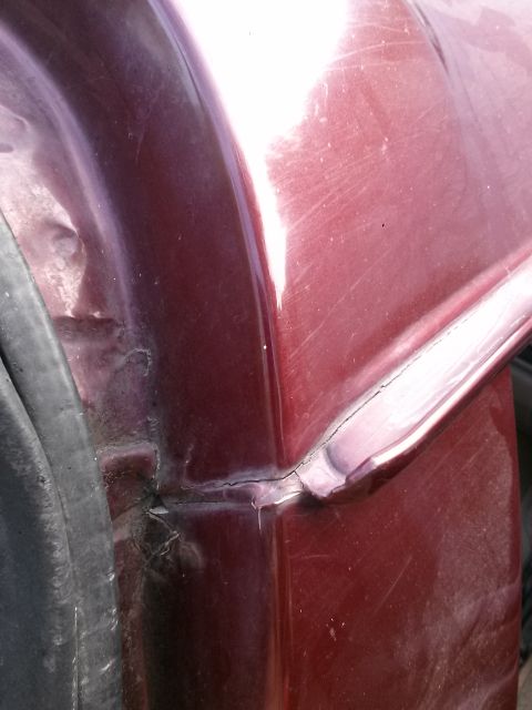 1996 Ford Bronco - Close-up view of the damaged maroon body panel of a vehicle, showing a cracked seam and denting near the wheel well.