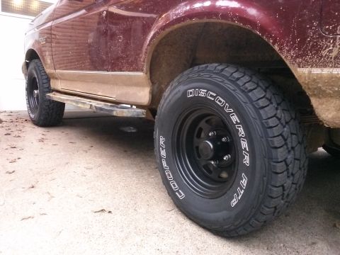 1996 Ford Bronco - A close-up, low-angle shot shows the rear passenger side of a muddy, maroon Ford Bronco. The vehicle features black aftermarket wheels with…